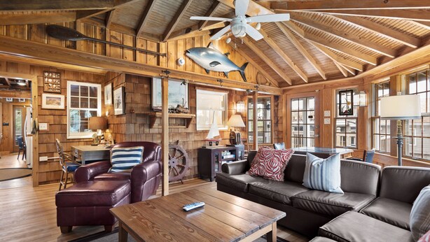 A cozy cabin interior with a wooden ceiling, a large ceiling fan, a leather sofa, a wooden coffee table, and a dining area with a table and chairs.