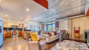 A spacious living area with a sofa, armchairs, and a rug. A kitchen with modern appliances and wooden cabinets is visible in the background. - Chic Condo Steps from Spokane River And Dining (Coeur d'Alene)