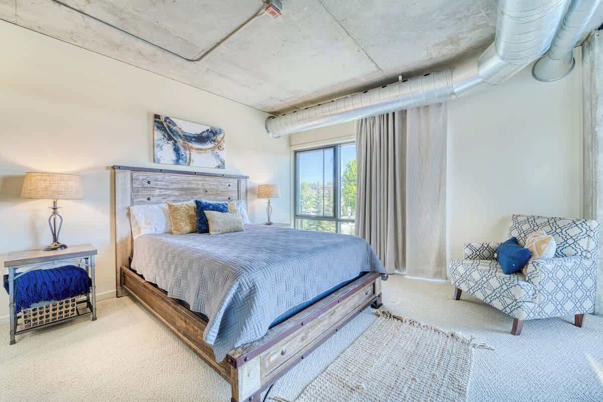 Un dormitorio con una cama de madera, dos mesitas de noche, un sillón estampado y un ventanal con cortinas.