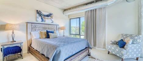Un dormitorio con una cama de madera, dos mesitas de noche, un sillón estampado y un ventanal con cortinas.