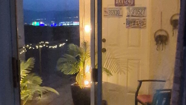 Vista de dentro de um quarto, com uma porta que dá para uma área externa. A porta possui uma placa com a inscrição 'LAS VEGAS STRIP'. Plantas e uma cadeira são visíveis do lado de fora.