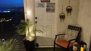 Uma varanda com porta, planta em vaso, duas cadeiras, uma mesa pequena e um lustre suspenso.