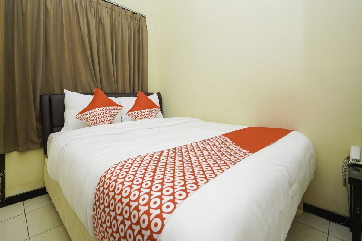A hotel room with a bed, brown curtains, white bedding, and orange pillows.