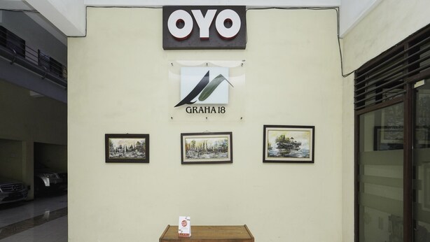 A building with a sign reading "OYO" and "GRAHA18". There are three framed pictures on the wall and a wooden table with a box on it. - Oyo 2902 Graha 18 (Surabaya)
