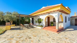Ein kleines, weißes Gebäude mit gelbem Dach und Steinböden. Es verfügt über eine überdachte Terrasse mit Tisch und Stühlen sowie einen kleinen Gartenbereich mit Blumentöpfen.