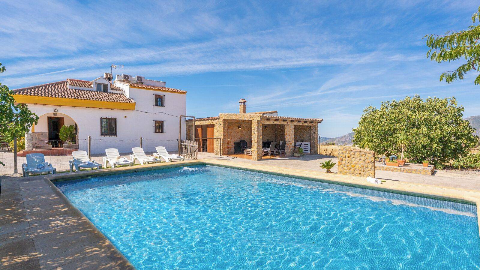 A swimming pool with lounge chairs and a stone structure, likely a spa or sauna, adjacent to a white building with a tiled roof.