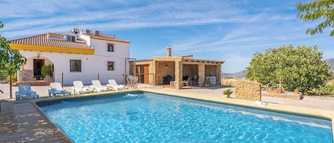 A swimming pool with lounge chairs and a stone structure, likely a spa or sauna, adjacent to a white building with a tiled roof.