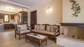 Ein Wohnzimmer mit Sofa, Sessel und Couchtisch. Rechts ist ein Kamin zu sehen, im Hintergrund befindet sich eine Küche.