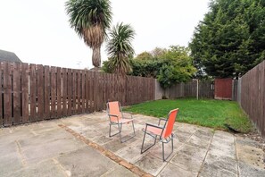 A patio with two metal chairs and a stone floor, surrounded by a wooden fence and greenery.