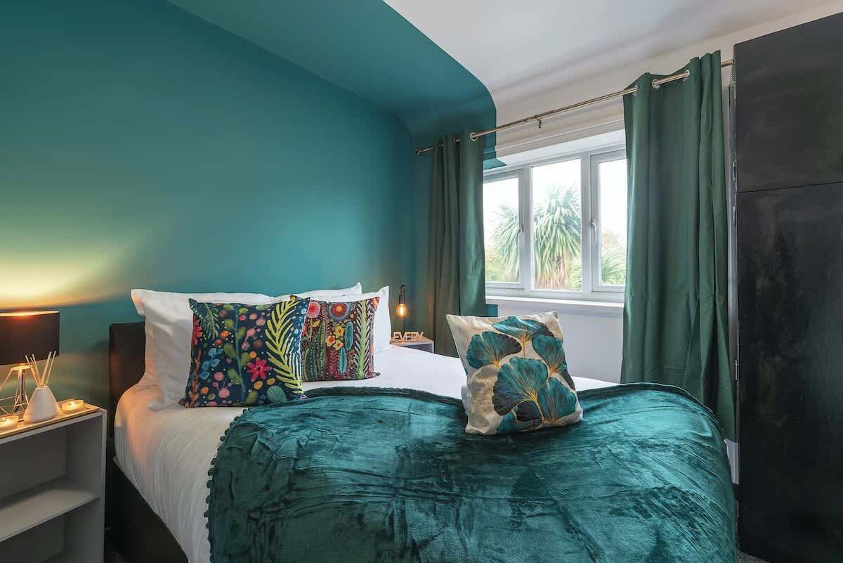 A bedroom with a bed, pillows, a nightstand, a window with curtains, and a view of palm trees outside.
