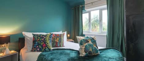 Un dormitorio con cama, almohadas, mesita de noche, ventana con cortinas y vistas a palmeras desde el exterior.