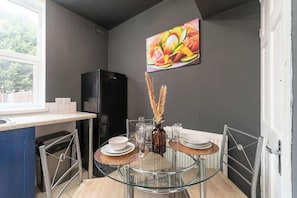A compact kitchen with a black refrigerator, a dining table with glass top, and a painting of fruits on the wall. - Spacious House, Garden, Private Parking, Near Hull Uni & Beverley (Kingston upon Hull)