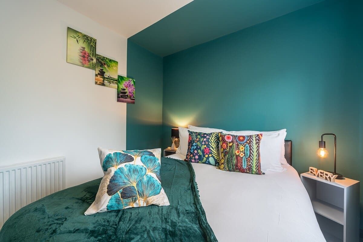 Une chambre avec un mur d’accent de couleur turquoise, un lit avec deux oreillers, une table de chevet avec une lampe et des œuvres d’art murales.