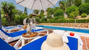 A poolside area with lounge chairs, a table with a bottle and glasses, and a stone wall in the background. - Villa Maroma Viñuela by Ruralidays (Málaga)