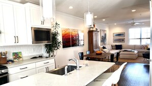Una cocina moderna con armarios blancos, una isla grande con taburetes de bar, un comedor con mesa y sillas, y una sala de estar con sofá y ventilador de techo.