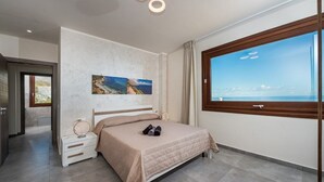 Un dormitorio con una cama, una mesita de noche, un ventanal con vistas al mar y una puerta que da a otra habitación.