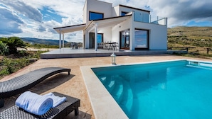 Una casa moderna de dos pisos con piscina, mobiliario exterior y vistas a las montañas.