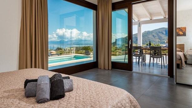 Habitación de hotel con una cama grande, vistas a una piscina y montañas, y un balcón con mesa y sillas.