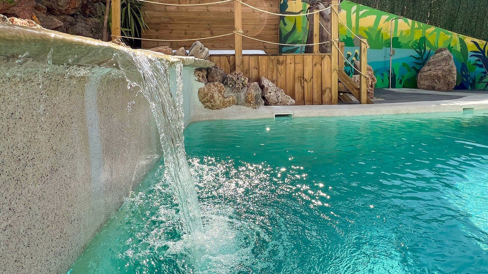 A swimming pool with a waterfall feature, surrounded by a wooden deck and a mural.