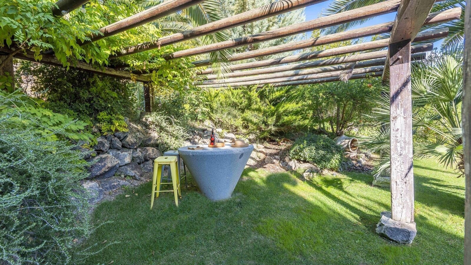 Een stenen tafel met een fles en een boek, omringd door groen en een houten pergola.