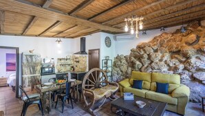 A rustic interior with a stone wall, wooden ceiling beams, a dining area with a wooden table and chairs, and a yellow sofa with blue cushions. - Casa de La  Cascada Iznájar by Ruralidays (Córdoba)