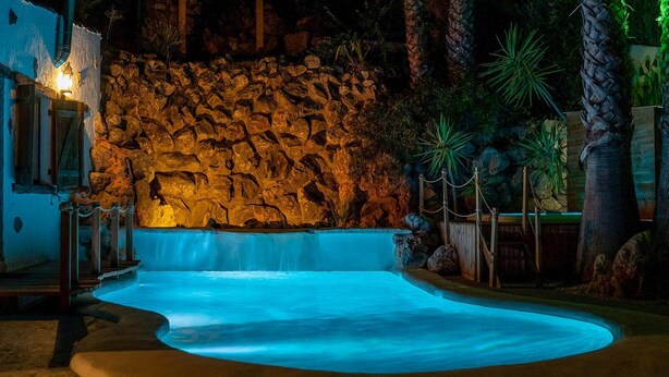 A swimming pool with a waterfall feature, surrounded by a stone wall and tropical plants. - Casa de La  Cascada Iznájar by Ruralidays (Córdoba)