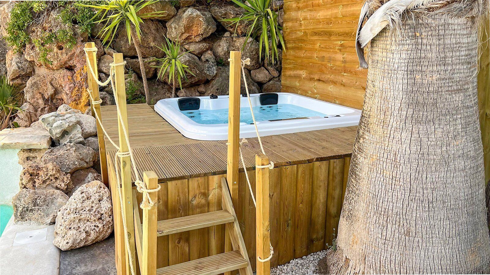 A hot tub with a wooden deck, surrounded by rocks and plants.