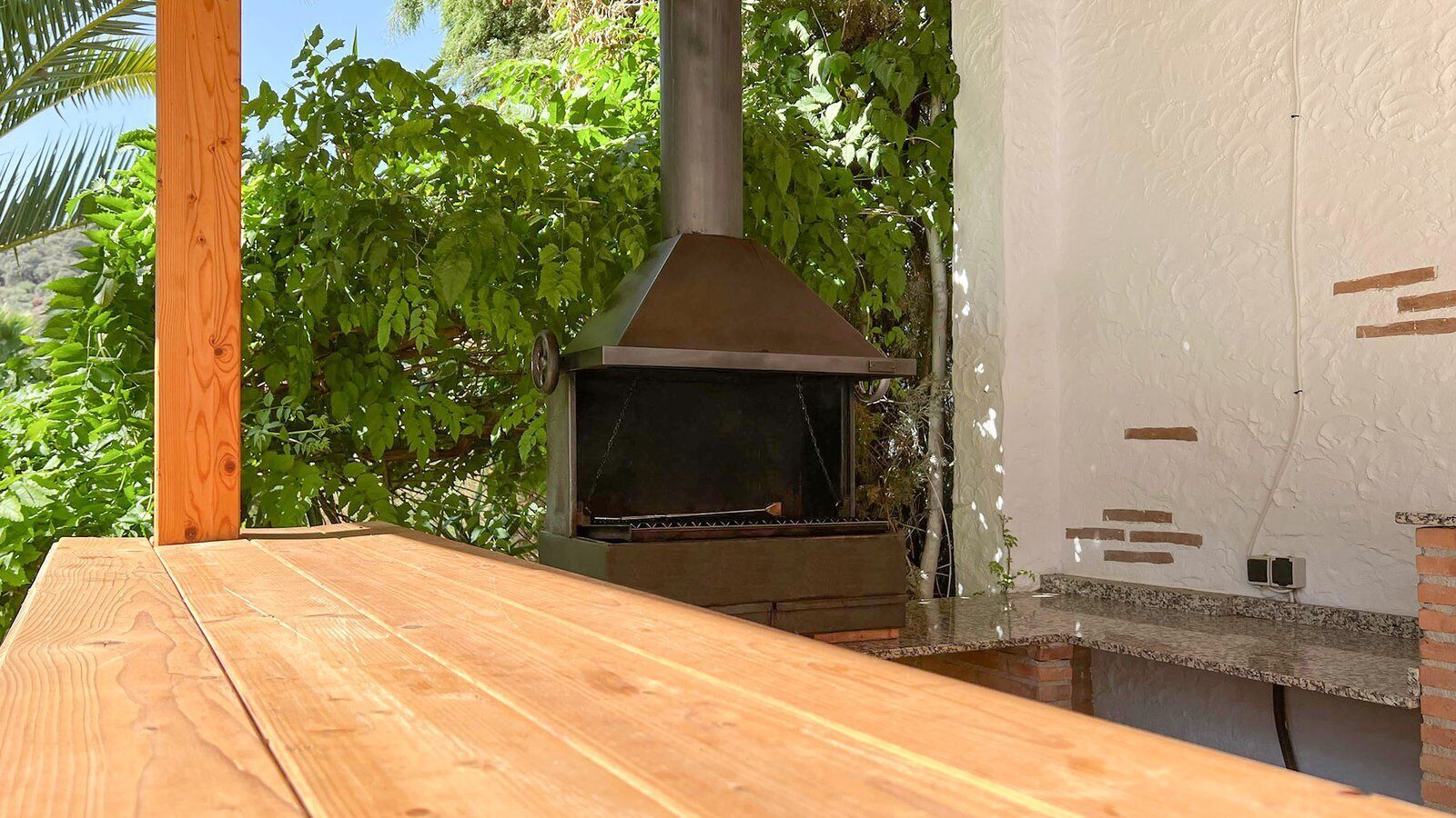 Een houten tuintafel met een barbecue van baksteen op de achtergrond.