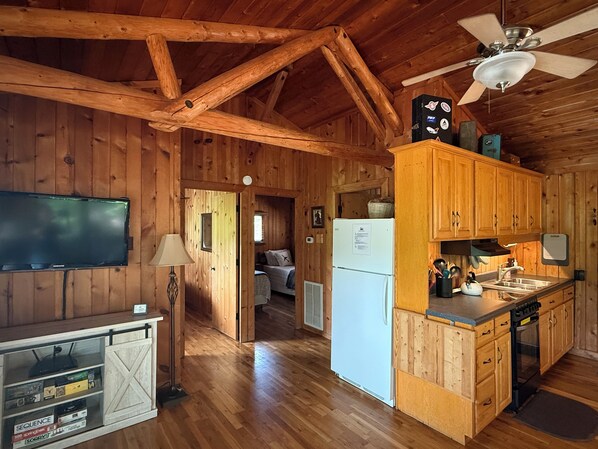 A rustic cabin interior with wooden walls, a ceiling fan, a kitchen with wooden cabinets, a refrigerator, and a television mounted on the wall.