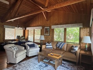 Interior acogedor de una cabaña con techo de madera, un sofá, dos sillones, una mesa de centro y un comedor con una mesa y sillas.