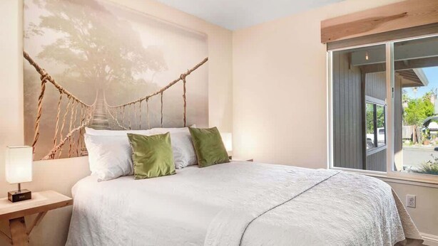 A bedroom with a large bed, a nightstand, a wall-mounted lamp, and a view of the outdoors through a window. - Sonoma Valley Vacation Home (Santa Rosa)