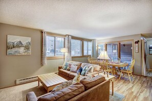 A cozy living room with a brown leather sofa, a wooden coffee table, and a dining table with chairs. There is a painting on the wall and a window with blinds.