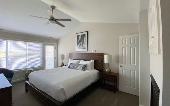 Une chambre à coucher comprenant un lit, deux tables de chevet, un ventilateur de plafond, une porte et une fenêtre avec des stores.