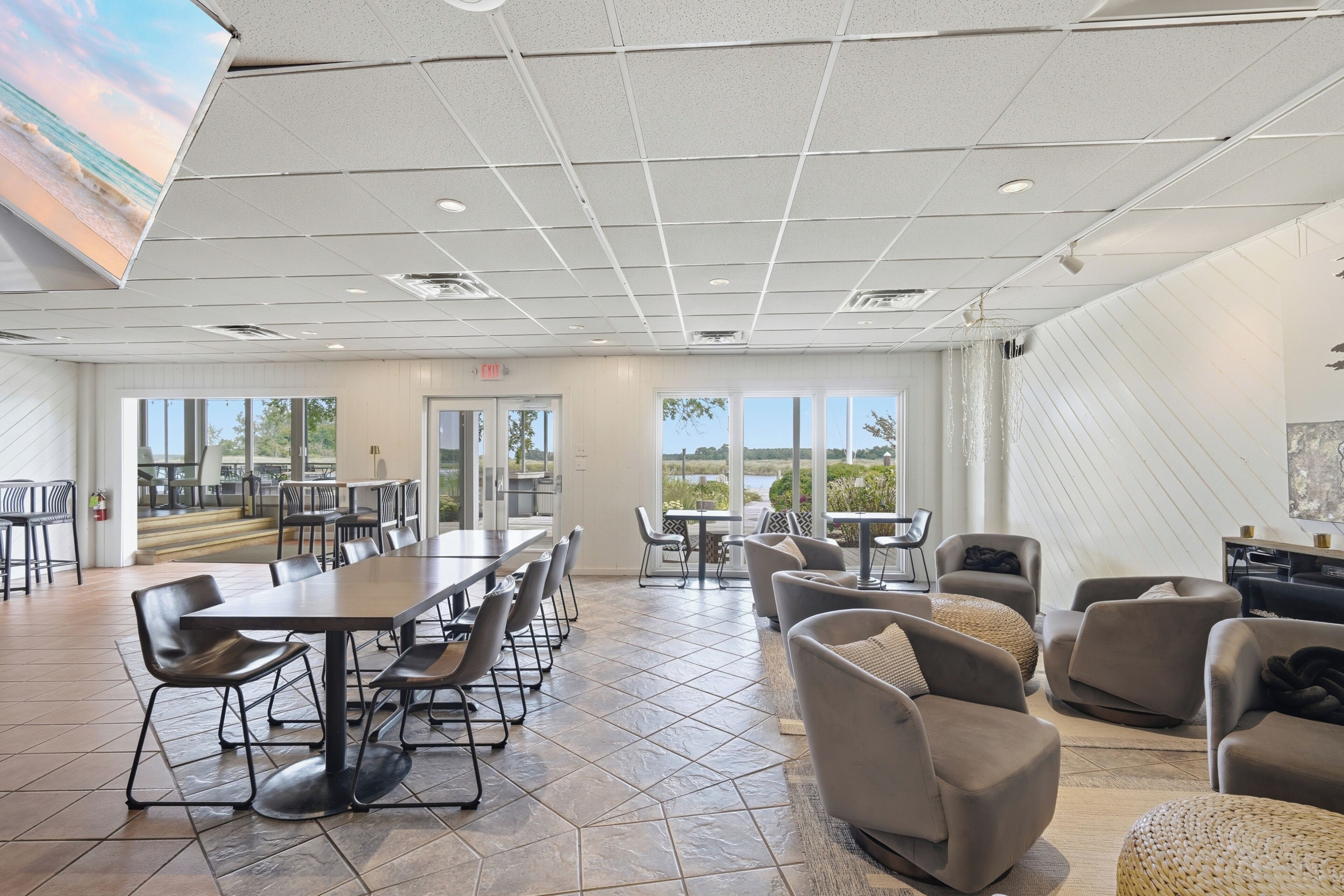 A spacious indoor area with a long table and chairs, a seating area with sofas, and large windows offering a view of the outdoors.