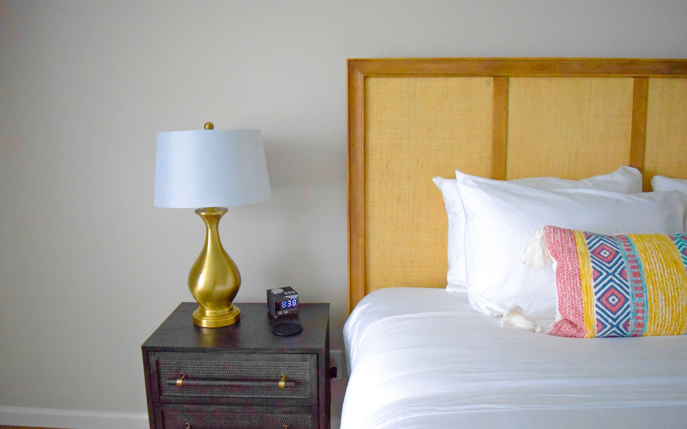 A neatly made bed with white linens and a colorful throw blanket, a bedside table with a lamp and a clock, and a wooden headboard.