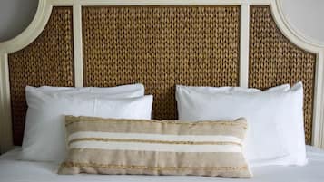 Una cama con ropa de cama blanca y dos almohadas, una a rayas y otra lisa, frente a un cabecero tejido.