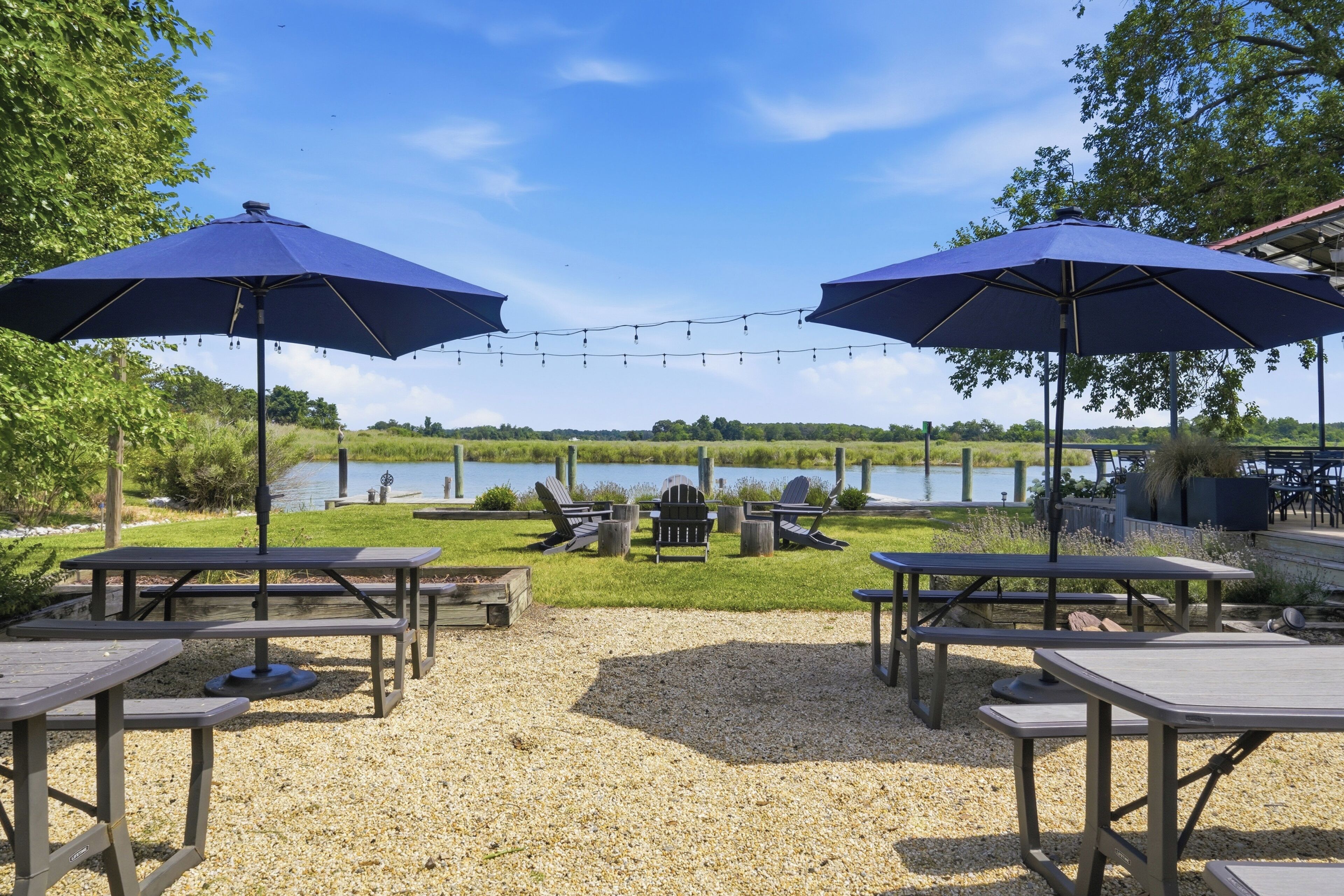 A lakeside patio with picnic tables, two large umbrellas, and lounge chairs.