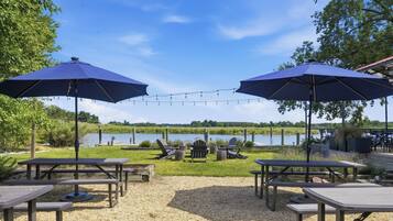 A lakeside patio with picnic tables, two large umbrellas, and lounge chairs.