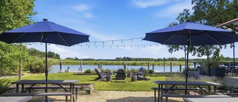 A lakeside patio with picnic tables, two large umbrellas, and lounge chairs.