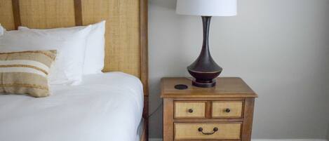 A neatly made bed with white linens and a wooden headboard, a bedside lamp on a wooden nightstand with drawers, and a plain wall in the background.