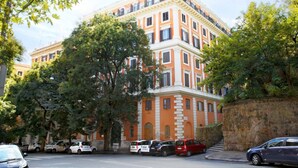 Ein mehrstöckiges Gebäude mit steinernem Sockel und orangener Fassade, umgeben von Bäumen und parkenden Autos.
