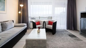 Un salon moderne avec un canapé gris, deux fauteuils avec des coussins rouges, une table basse ornée d’un vase et de grandes fenêtres avec des rideaux légers.