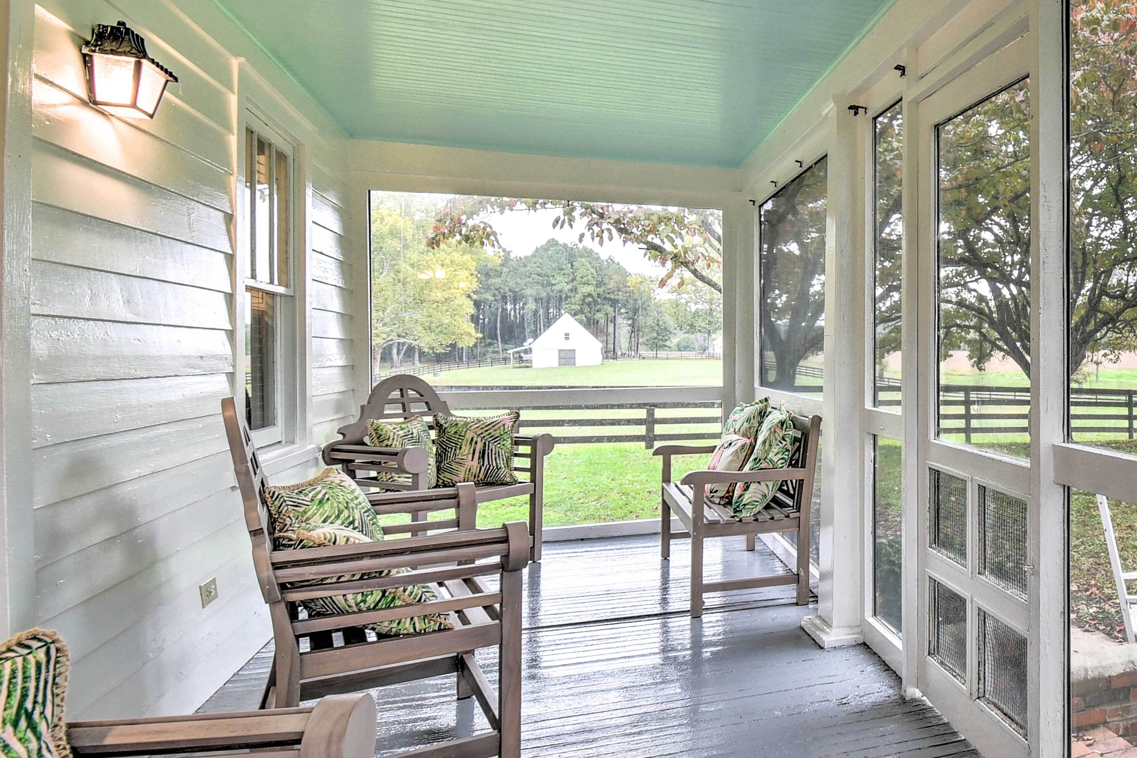 Eine Veranda mit Holzstühlen und einer Bank, einem weißen Geländer und Blick auf eine grüne Fläche mit Bäumen und einem Gebäude.