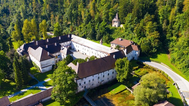 An aerial view of a historic building complex surrounded by dense forest, featuring multiple buildings with distinct architectural styles, a central courtyard, and a small tower.