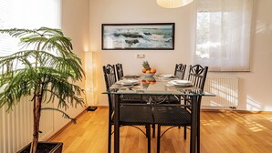 A dining area with a glass table, black chairs, a potted plant, a framed picture on the wall, and a window with curtains.