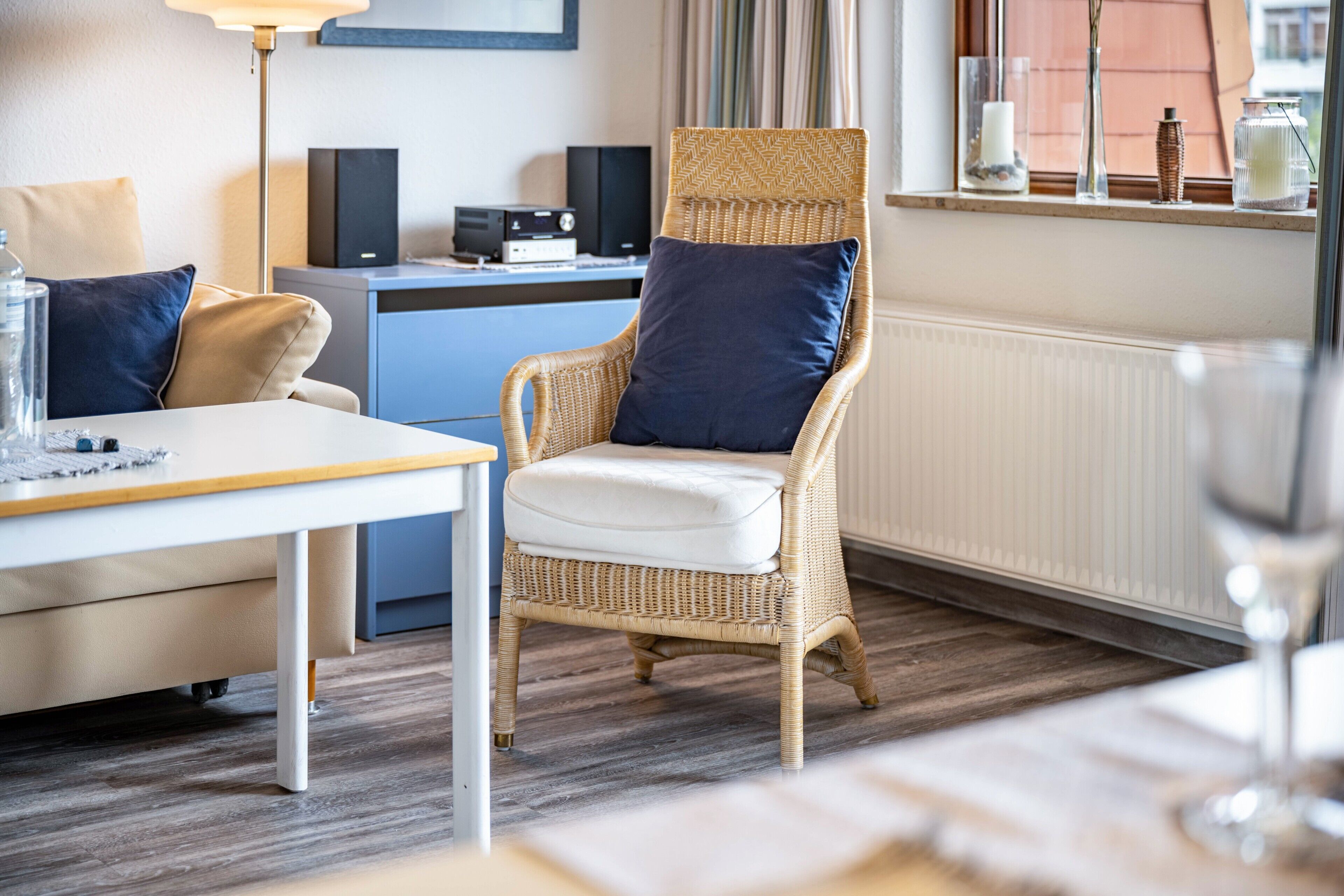 Een kamer met een rieten stoel, een tafel met kussen, een bureau met computer en een raam met gordijnen.