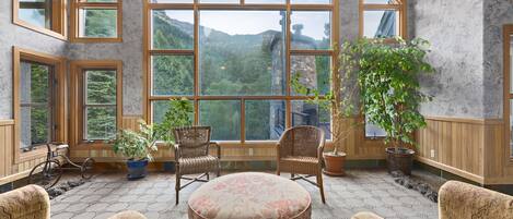 Une pièce chaleureuse avec de grandes fenêtres, des fauteuils en osier, un pouf à motifs floraux et des plantes en pot.
