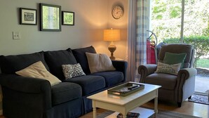 A living room with a dark blue sofa, a white coffee table, a lamp, a clock, and a chair with a cushion. - E13 Build family memories,  comfortable, clean (St. Simons Island)