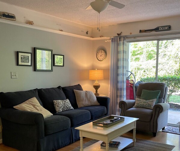 A living room with a dark blue sofa, a white coffee table, a side table with a lamp, a clock on the wall, and a view of the outdoors through a glass door.