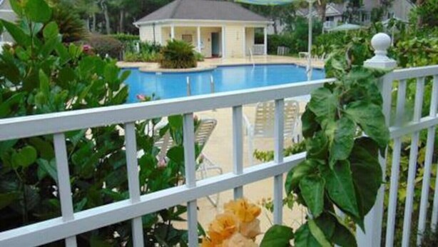 A pool area with a small building, surrounded by lush greenery and a white fence. - E13 Build family memories,  comfortable, clean (St. Simons Island)